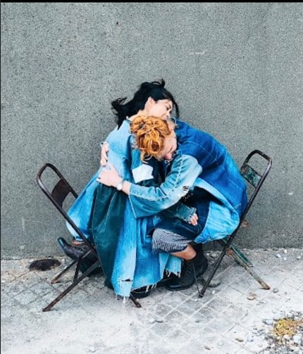 Two people wearing Maja's designs embrace while sitting on two facing chairs